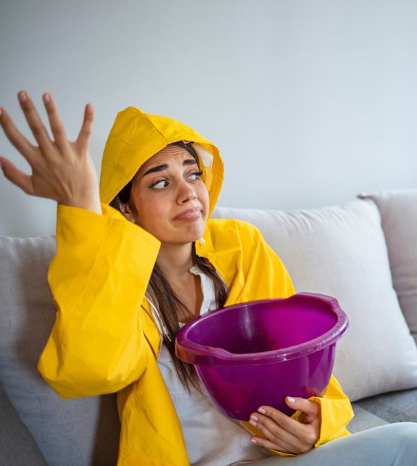 Woman on couch holding pail to catch water dripping from ceiling